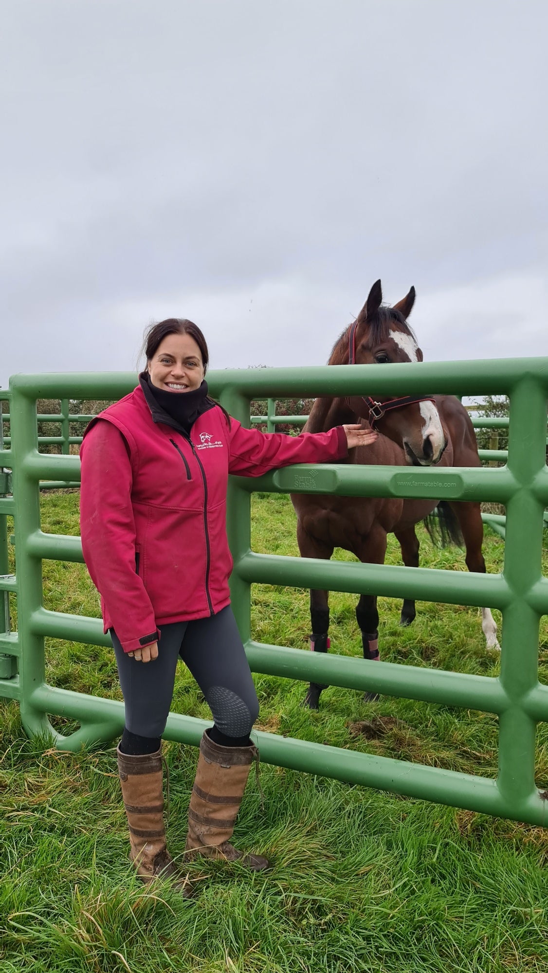 Ruffnek black fleece neck warmer neck gaiter shown worn by a horse woman stood next to a horse in a livery yard.